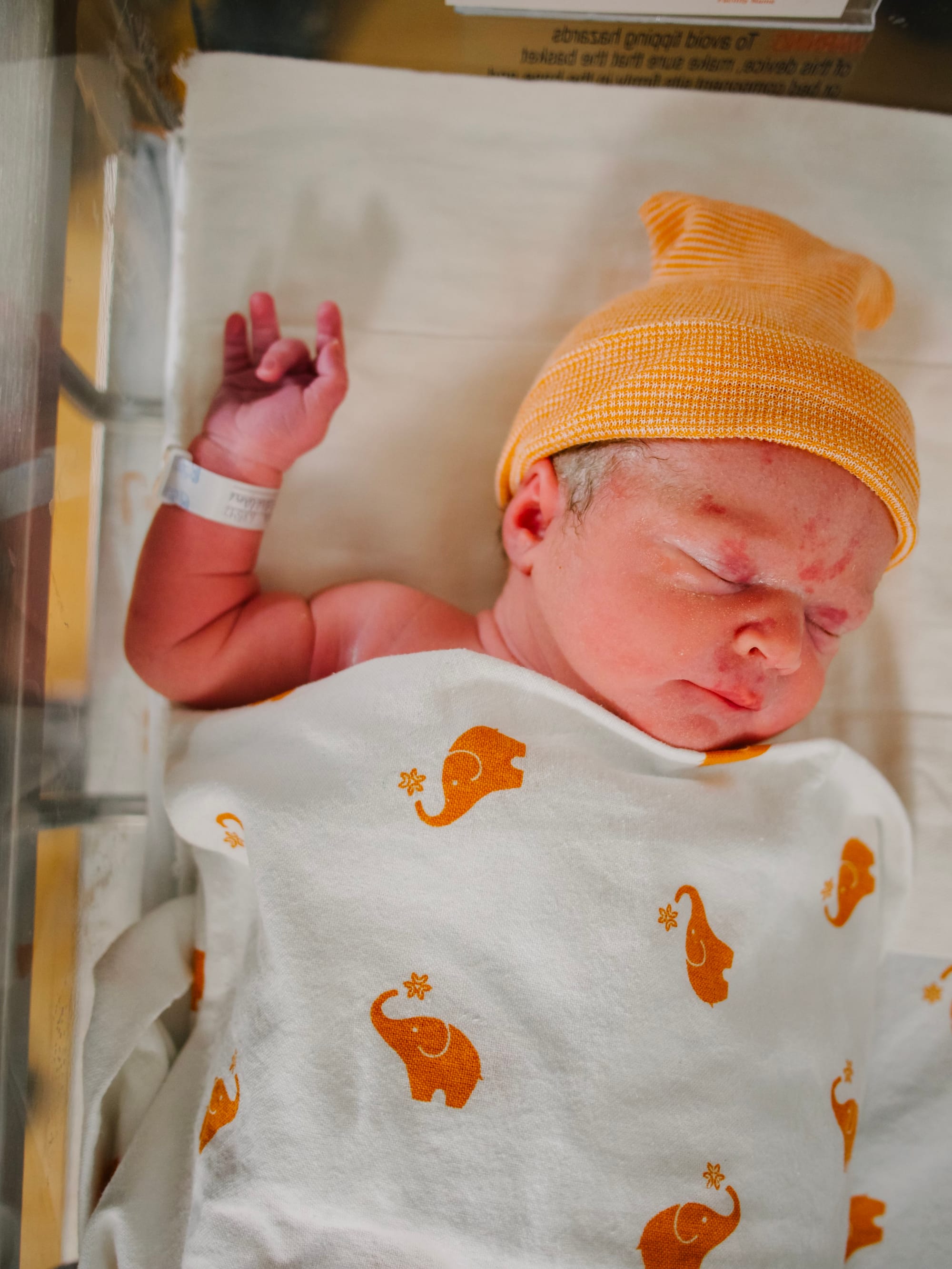 Photo of a baby in a hospital bassinet. Her hand almost forms a rock-on sign.