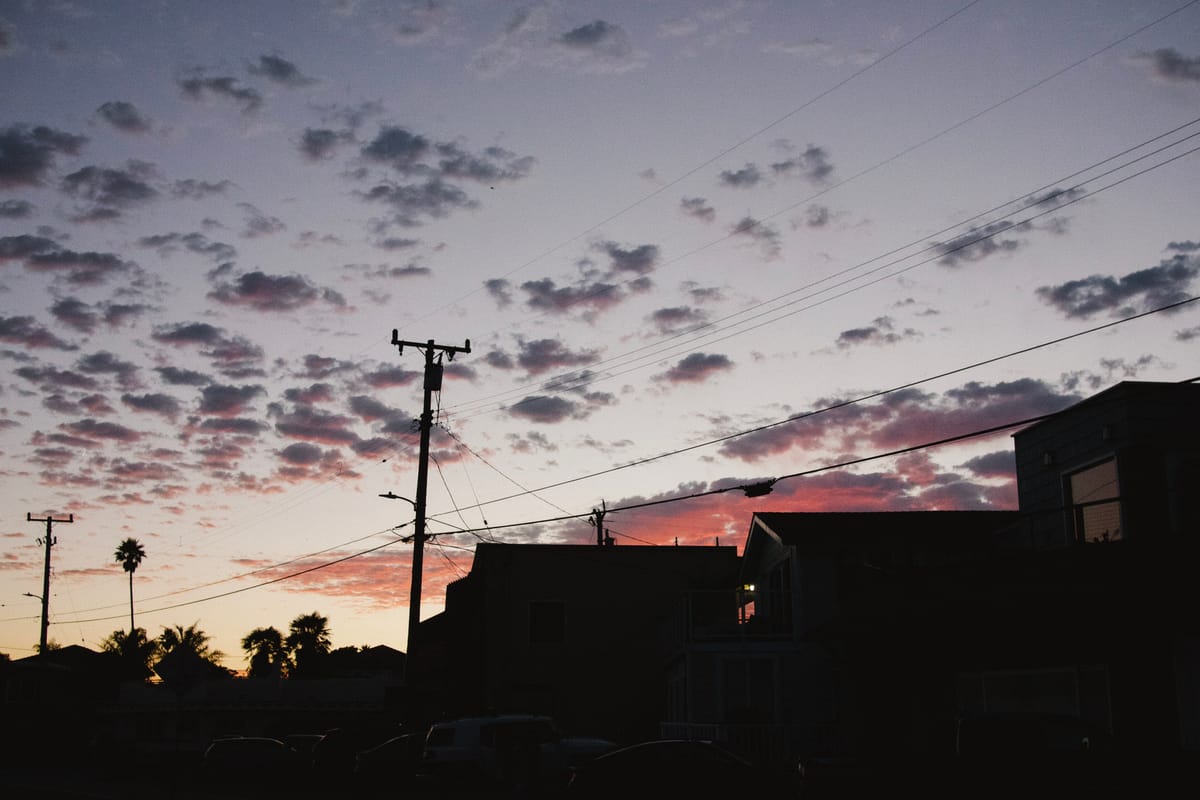 Photo of a sunset. Sparse clouds highlighted in pink dot the sky. Houses, palm trees, and telephone poles line the bottom of the frame.