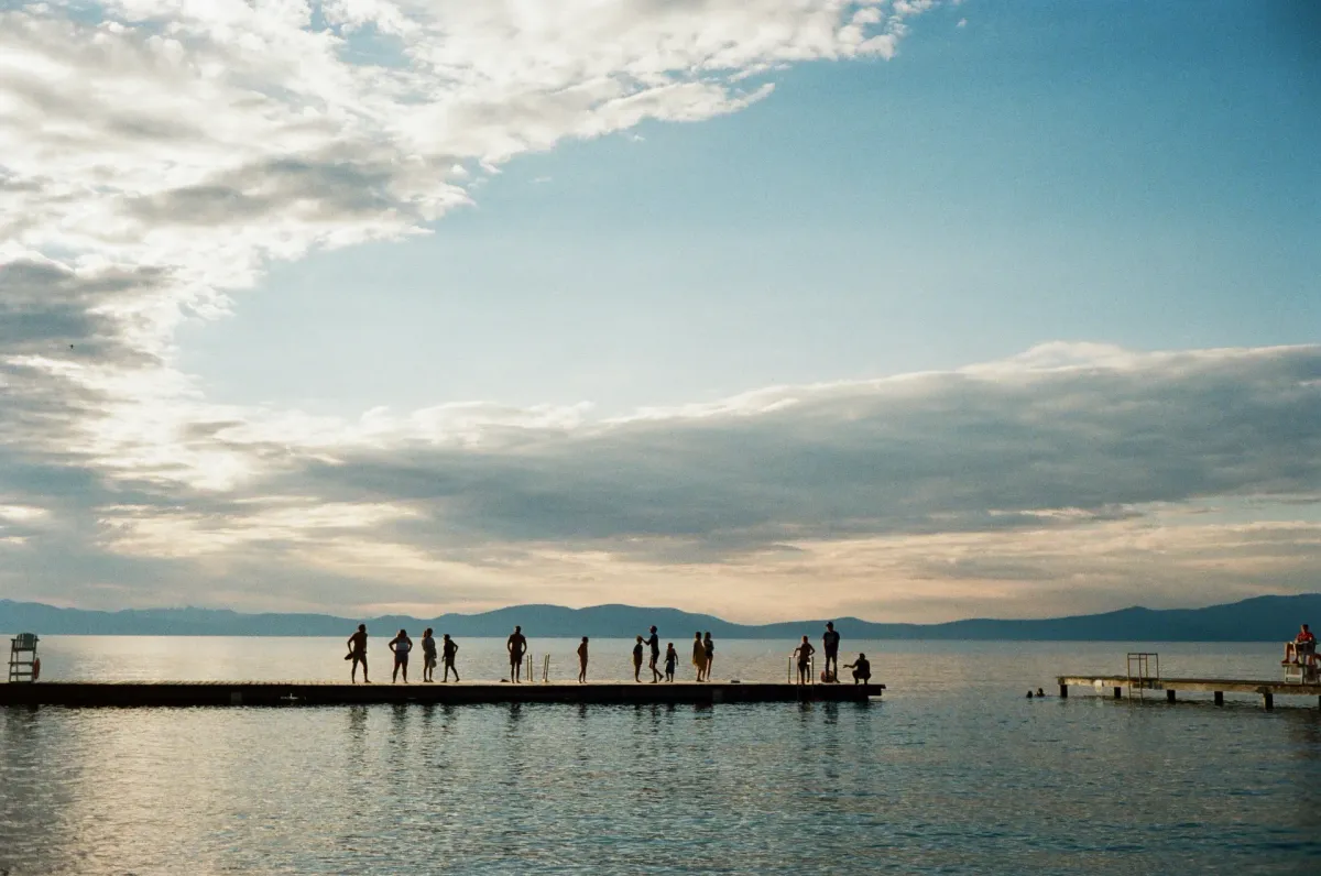 Film photo. A group of humans stand on a dock on Lake Tahoe. The sun sets behind them, across the lake.